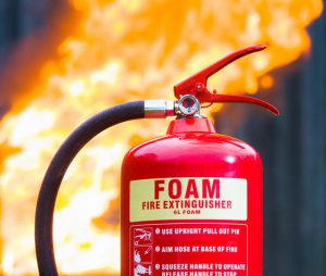 Close-up of a foam based fire extinguisher ready for use, with the flames of a large fire in the background.